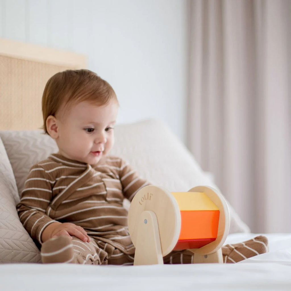 A baby sits on a bed playing happily with Totli - The Spinning Drum